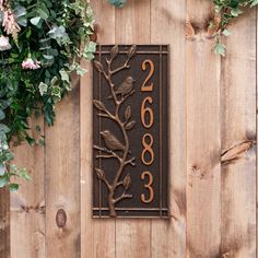 a house number sign on a wooden fence with flowers and greenery around the door