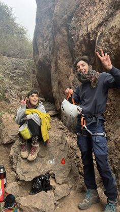 two people standing next to each other in front of some rocks and trees with one person holding up the peace sign