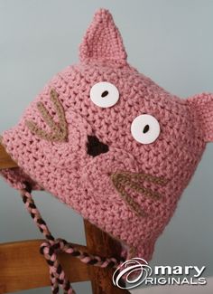 a crocheted pink cat hat with eyes and ears on top of a wooden chair