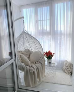 a white hanging chair next to a window with sheer curtains and flowers in the corner