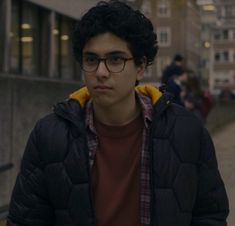 a young man wearing glasses standing in front of a building with people walking on the sidewalk behind him