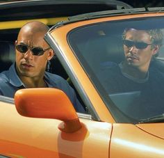 two men sitting in the drivers seat of a sports car, one with sunglasses on