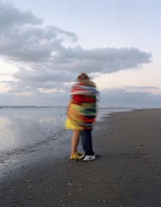 a man and woman hugging on the beach with clouds in the sky behind them at sunset