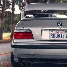 the back end of a silver bmw car parked on the side of the road with trees in the background