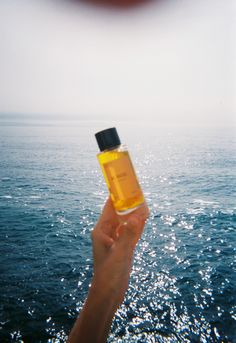 a hand holding up a bottle of oil over the ocean with water in the background