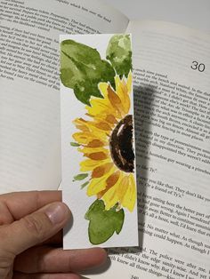 a hand holding an open book with a watercolor painting of a sunflower on it