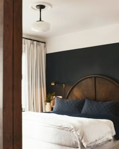 a bed with white sheets and blue pillows in a dark colored room, next to a window