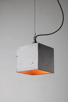 a concrete light fixture hanging from a wire with an orange light in the center and white wall behind it