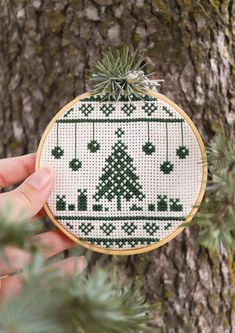 a hand holding up a cross stitch christmas ornament in front of a tree