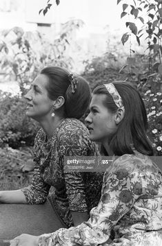 two women sitting next to each other on a bench in front of some bushes and flowers