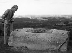 a man standing next to a large rock in the middle of a field with a sign on it