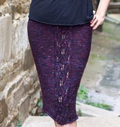 a woman in black shirt and purple skirt standing next to brick wall with her hand on her hip