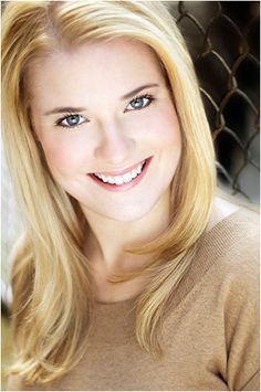 a woman with blonde hair and blue eyes smiles at the camera in front of a chain link fence