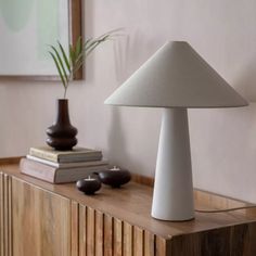 a white table lamp sitting on top of a wooden dresser next to a plant and books