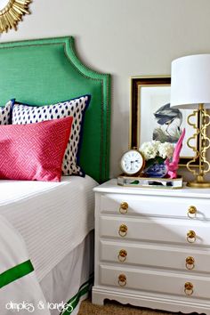 a white bed topped with lots of pillows next to a green headboard and dresser