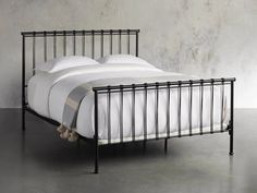 an iron bed frame with white sheets and pillows on it, in front of a concrete wall