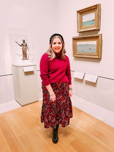 a woman in a red sweater and black floral skirt standing in front of some paintings