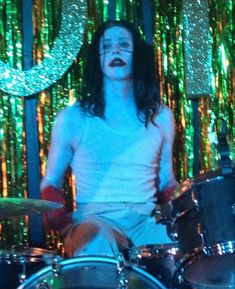 a man sitting on top of a drum set in front of a green curtained background