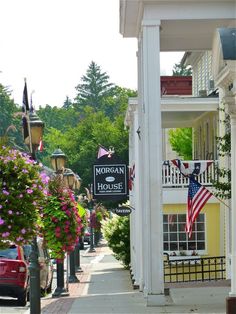 an american flag hanging from the side of a white building on a street with flowers in front of it