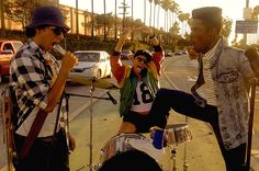 two men are playing drums and singing on the street while another man holds his leg up in the air