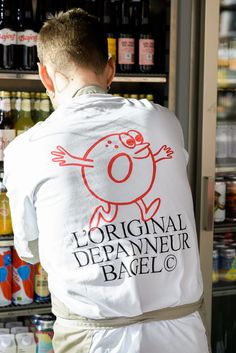 a man standing in front of a shelf filled with bottles and cans, wearing a white t - shirt that says original depanneur bagelc