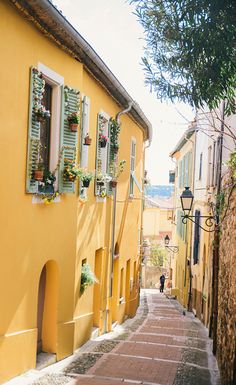 a narrow street with yellow buildings and green shutters on the windows, lined with potted plants