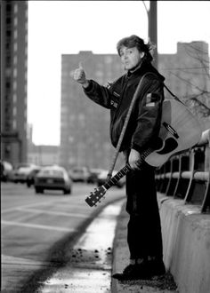 a woman standing on the side of a road holding a guitar