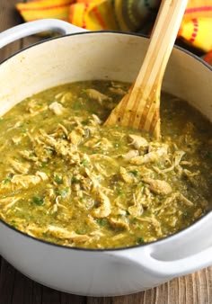 a wooden spoon in a white pot filled with green soup and chicken meat, on a wood table