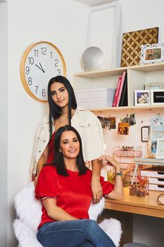 two women sitting on a chair in front of a desk with a clock above them