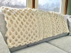 a white knitted pillow sitting on top of a gray couch next to a window