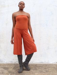 a woman standing in front of a white wall wearing an orange top and brown pants