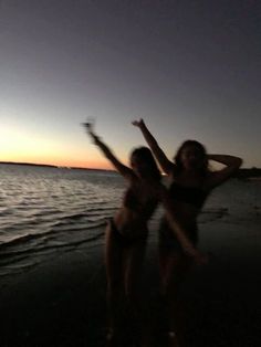 two women in bathing suits standing on the beach at night with their arms raised up