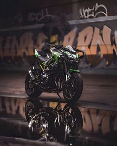 a green and black motorcycle parked in front of a graffiti covered wall with its reflection on the ground