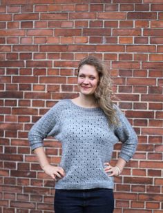 a woman standing in front of a brick wall wearing black pants and a gray sweater