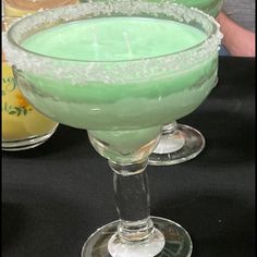 two margaritas sitting next to each other on top of a black table with drinks in the background