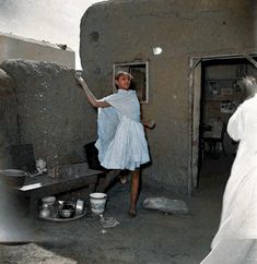 a woman in a blue dress is standing near a stove and some pots on the ground