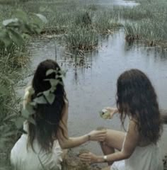 two young women sitting on the ground in front of a body of water and plants