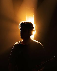 a man standing in front of a light with his head turned to the side while holding a guitar