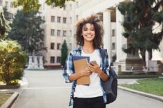 a woman walking down the street with a backpack and notebook in her hand while holding a cell phone