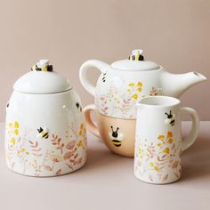 three teapots and two cups with bees painted on them, sitting next to each other