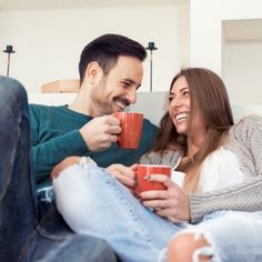 a man and woman sitting on a couch holding coffee mugs, smiling at each other