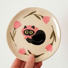 a hand holding a plate with a black cat on it's face and pink flowers