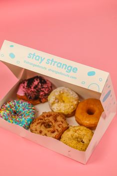 an open box of donuts with sprinkles on pink paper and blue lettering that says stay strange