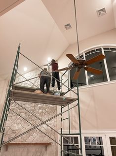 two men on scaffolding working on a ceiling fan in a room with stone walls