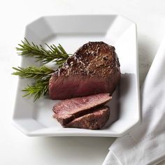 a piece of steak on a white plate with a sprig of rosemary next to it