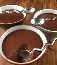 three bowls filled with chocolate pudding on top of a wooden table next to two spoons