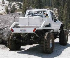 a white truck parked on top of a snow covered slope next to rocks and trees