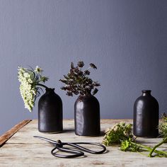 three black vases sitting on top of a wooden table next to flowers and scissors