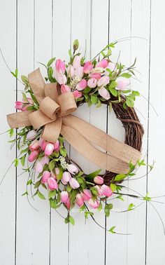 a wreath with pink flowers and green leaves on a white wooden wall, tied with a jute bow