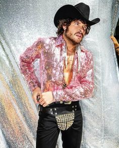 a man in a pink shirt and black pants standing next to a silver curtain with his hands on his hips